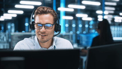 a guy smirking with a headset on, looking at some monitors in a room with like 20 fluorescent tubes lighting it, way more than is necessary